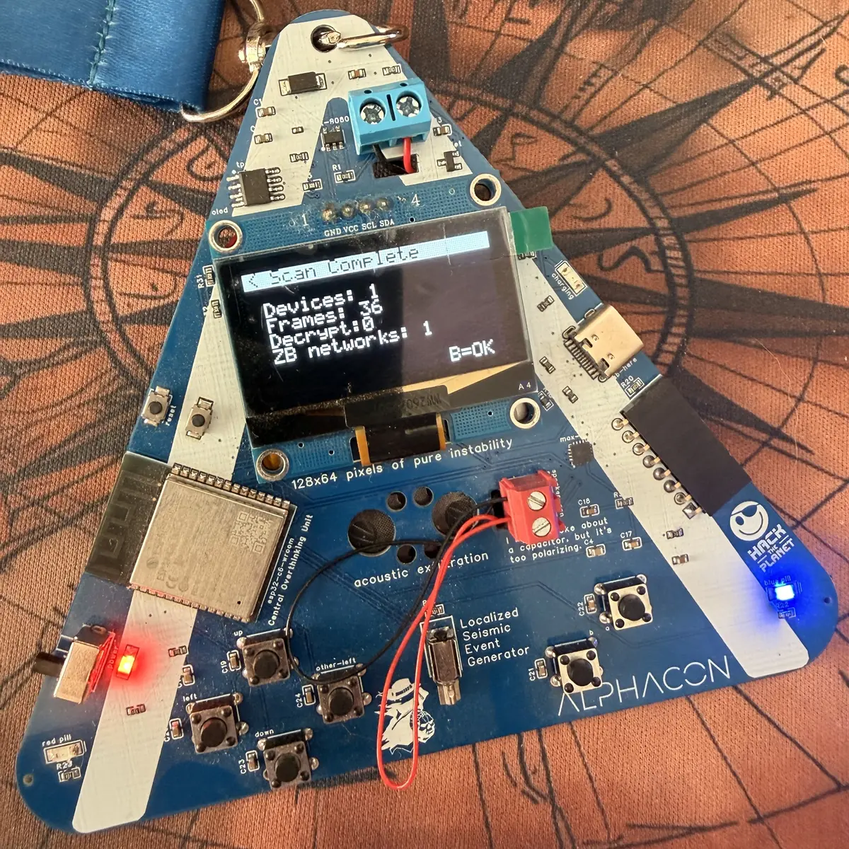 Przód badge'a Alphacon — trójkątna płytka drukowana z wyświetlaczem OLED i modułem ESP32-C6